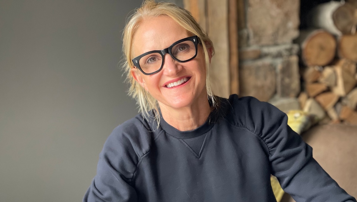 Mel Robbins smiling in front on stone fireplace wearing blue sweater and black glasses